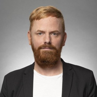 Mann mit roten Haaren und Vollbart, der ein weißes T-Shirt und eine schwarze Anzugjacke trägt